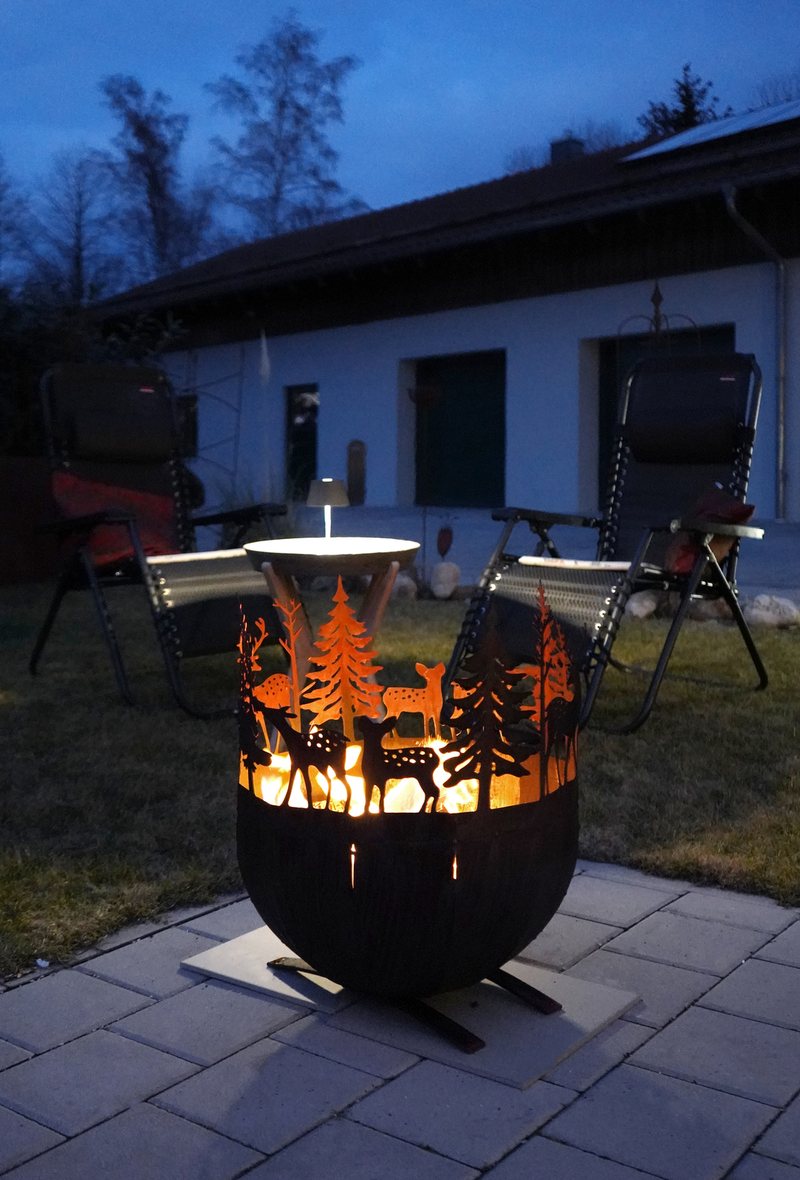 Feuerstelle mit Hirsch- und Baummotiv, brennend auf einer Terrasse in der Abenddämmerung