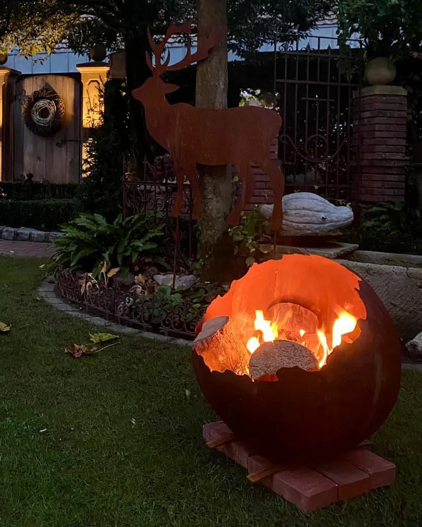 Feuerkugel aus Stahl mit brennendem Holz, dahinter eine Hirsch-Gartenfigur aus Stahl, im abendlichen Garten