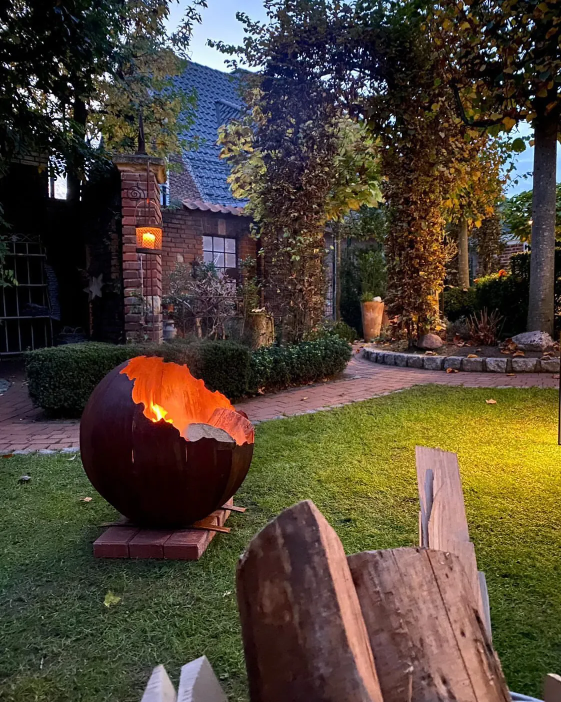 Feuerkugel aus Stahl mit brennendem Holz vor einem Haus mit Laterne, am Abend